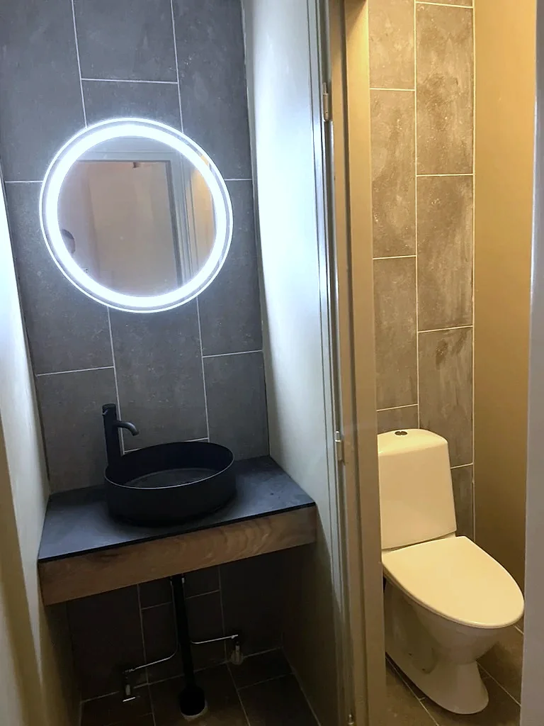 Hygienutrymme med mörkt kakel, runt handfat och belyst spegel
